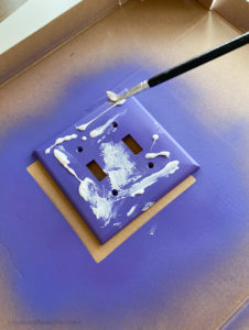 Painting glue on the purple switch plate cover before applying the glitter