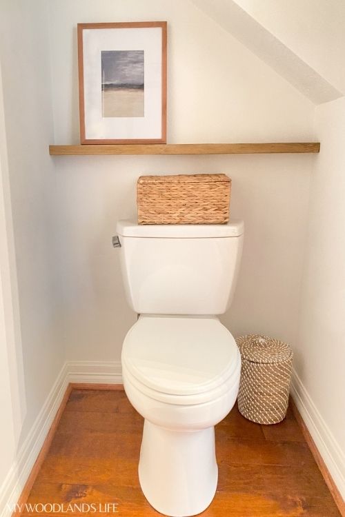 Small powder room with toilet, basket on top of toilet and wooden shelf on wall behind toilet 