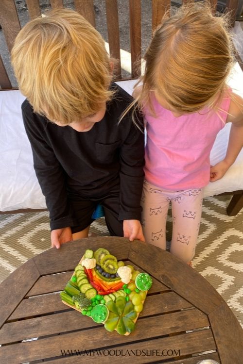 Two children looking at fruit and veggie snack board on wood table