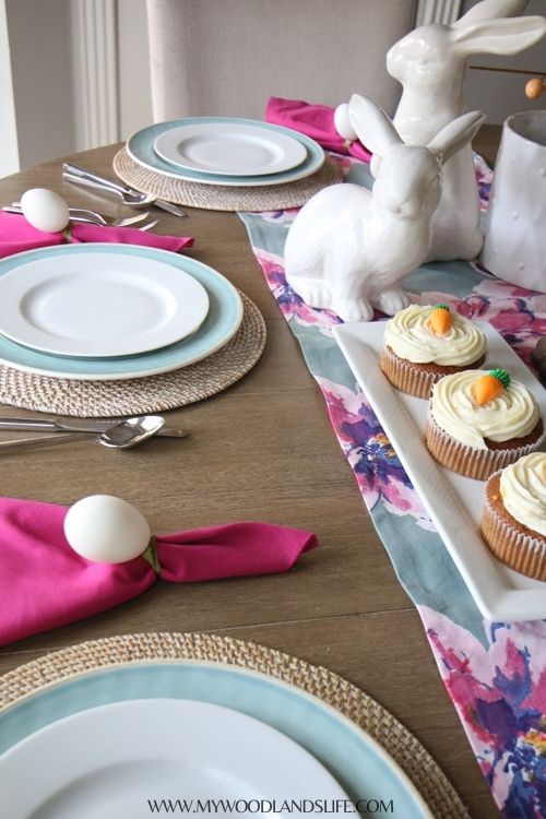 Easter themed table setting with blue and pink floral table runner, white ceramic rabbits, mini carrot cakes on white serving tray, hot pink napkins and blue and white plates on raffia chargers